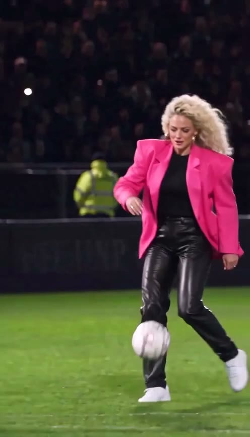 A goalkeeper dives past the woman in a pink blazer